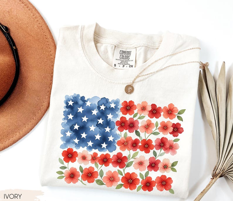 May include: Ivory-colored t-shirt featuring a watercolor design of the American flag, with blue stars and red and pink flowers. The shirt has a crew neck and short sleeves. A gold necklace with a pendant is draped across the shirt.