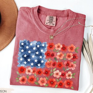 May include: Crimson-colored t-shirt featuring a floral American flag design. The flag's stars are white, the field is blue, and the stripes are represented by red and pink flowers. The shirt has a crew neck and short sleeves.