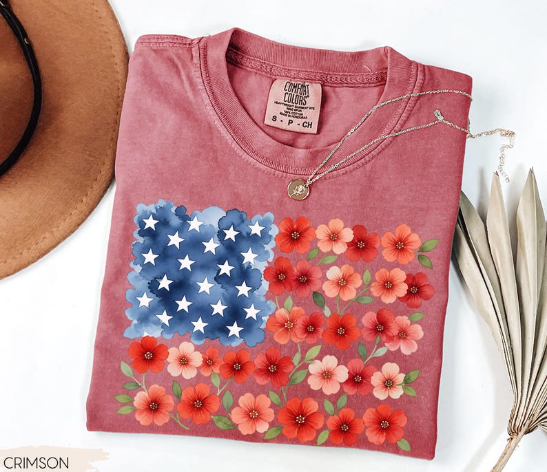 May include: Crimson-colored t-shirt featuring a floral American flag design. The flag's stars are white, the field is blue, and the stripes are represented by red and pink flowers. The shirt has a crew neck and short sleeves.