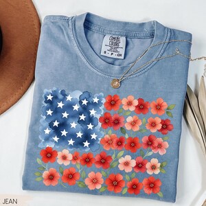 May include: Blue jean colored t-shirt with a floral American flag design. The flag features a watercolor-style blue field with white stars and red and pink flowers. The shirt has a Comfort Colors tag.