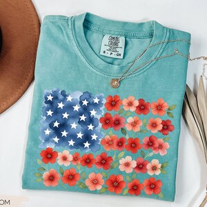 May include: A turquoise Comfort Colors t-shirt featuring a floral American flag design. The flag design includes a watercolor blue square with white stars and red and pink flowers. A gold necklace is draped on the shirt.