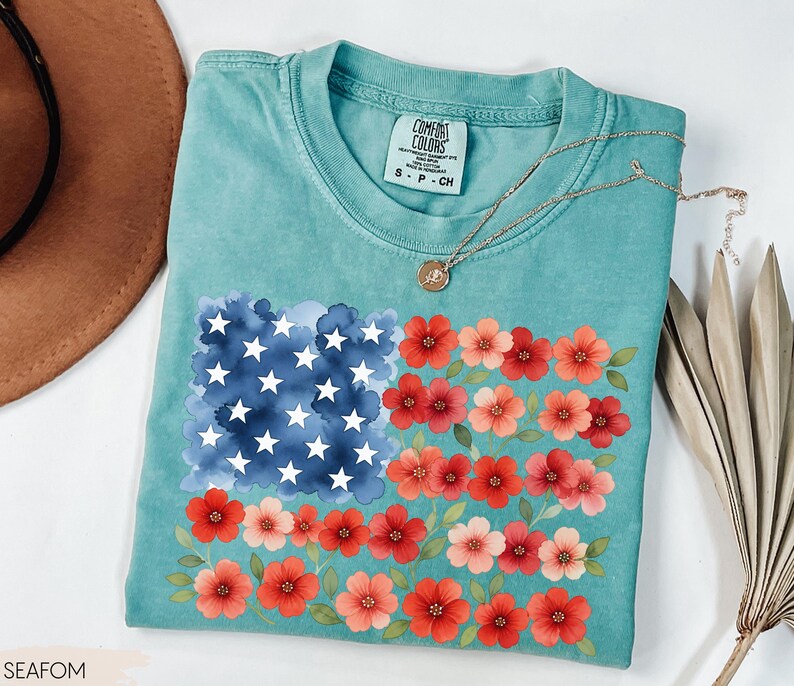 May include: A turquoise Comfort Colors t-shirt featuring a floral American flag design. The flag design includes a watercolor blue square with white stars and red and pink flowers. A gold necklace is draped on the shirt.