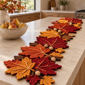 Pode incluir: Um caminho de mesa em crochê com um padrão repetitivo de folhas de bordo de outono em tons de vermelho, laranja e castanho, acentuado com pequenas bolotas de crochê. As folhas têm uma textura visível e estão dispostas numa fila longa.