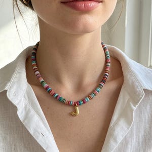 Pode incluir: Um colar com um fio de pequenas contas multicoloridas em forma de disco em tons de rosa, azul, verde e laranja. Um pingente de metal dourado brilhante em forma de letra maiúscula J está pendurado no centro do colar de contas.
