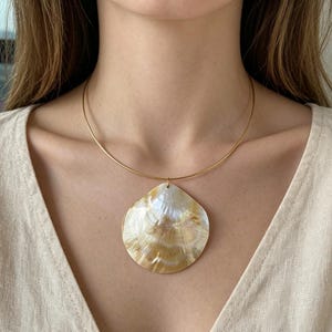 May include: A thin gold-toned metal choker necklace featuring a large, iridescent seashell pendant. The shell displays natural shades of cream, white, and golden-brown with a smooth, polished finish, suspended from a simple, circular wire band.