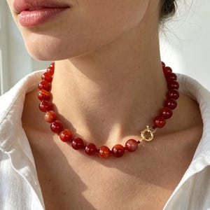 May include: A necklace featuring smooth, round, deep orange-red carnelian beads strung together. The necklace is secured with a circular gold-toned spring ring clasp positioned on the side, connecting the beaded strand to complete the circular design.