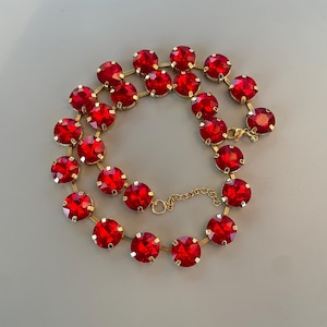 Peut inclure: Un collier doré composé d'une série de cristaux rouges ronds et facettés, sertis individuellement dans des griffes en métal. Le collier est disposé en cercle et comprend une chaînette d'extension dorée avec un fermoir mousqueton.