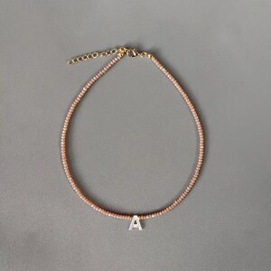 Puede incluir: Un collar de cuentas con pequeñas cuentas facetadas de color melocotón claro ensartadas en un cordón. El collar incluye un dije central con la letra A en color blanco y un cierre de mosquetón de metal dorado con una cadena de extensión.