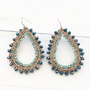 May include: A pair of teardrop-shaped earrings featuring a tan crocheted thread base adorned with faceted beads in shades of dark blue, teal, and light green. Each earring includes a silver-toned metal hook for attachment to the earlobe.