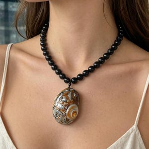 Può includere: Una collana di perline nere con un grande ciondolo ovale. Il ciondolo presenta un motivo a spirale di tonalità marroni, bianche e arancioni. La collana è indossata intorno al collo.