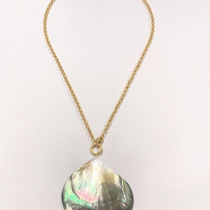 Pode incluir: Um colar de corrente dourada com um pingente grande e iridescente em forma de gota. O pingente tem um brilho perolado com tons de verde, rosa e prata. O colar é exibido em um fundo branco.