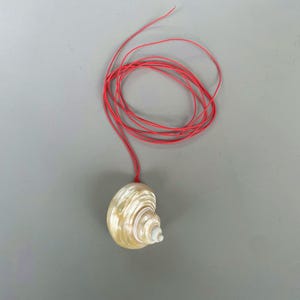 May include: A necklace featuring a polished, cream-colored spiral seashell pendant suspended from a thin, vibrant red cord. The shell exhibits a smooth, pearlescent sheen with subtle tan and white banding, while the red cord is coiled above.