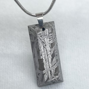 May include: A rectangular pendant made of a patterned, metallic material, suspended from a silver chain. The pendant has a unique, textured surface with intricate lines and patterns. The chain is a thin, silver snake chain.