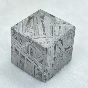 Puede incluir: Un cubo metálico con un patrón de Widmanstätten distintivo de bandas cristalinas plateadas y grises que se cruzan. El objeto geométrico muestra una textura superficial compleja y grabada con bordes afilados y un acabado pulido y brillante.
