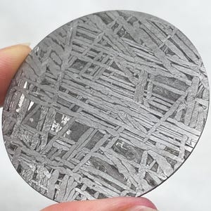 May include: A circular slice of iron meteorite featuring a distinct Widmanstätten pattern of intersecting metallic bands. The surface displays a complex, geometric grid of light gray crystalline structures contrasted against darker, recessed areas.