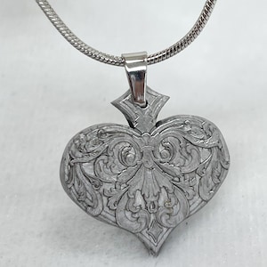 Può includere: Un ciondolo color argento a forma di cuore, finemente inciso con motivi floreali e a volute. Il ciondolo è sospeso a una catena d'argento. Il design è dettagliato e ornato, con un'estetica vintage.
