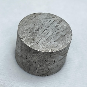 May include: A cylindrical, grey metallic object with a distinctive patterned surface. The pattern is made up of intersecting lines and shapes, creating a unique texture. The object is a solid, compact form, possibly a decorative or collectible item.
