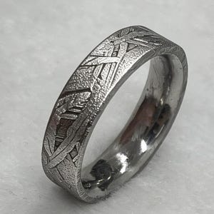 Könnte beinhalten: Ein silberfarbener Metallring mit einem ausgeprägten Widmanstätten-Muster auf der Außenfläche. Das Band hat eine texturierte, geätzte Oberfläche mit sich kreuzenden geometrischen Linien und eine glatte, polierte Innenseite, die Licht reflektiert.