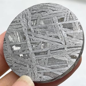 May include: A circular slice of iron meteorite featuring a distinct Widmanstätten pattern of intersecting metallic bands in shades of silver and dark grey. The surface displays a complex, crystalline geometric texture with varied light reflection.