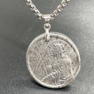 Può includere: Collana color argento con un ciondolo rotondo con un'immagine incisa dettagliata di una figura religiosa. Il ciondolo è attaccato a una catena. Il ciondolo ha un aspetto strutturato e metallico.