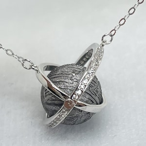 Puede incluir: Un collar de tono plateado con un colgante esférico de superficie oscura, texturizada y con cráteres. La esfera está rodeada por un anillo de plata pulida engastado con pequeñas piedras claras y brillantes, suspendido de una cadena fina.
