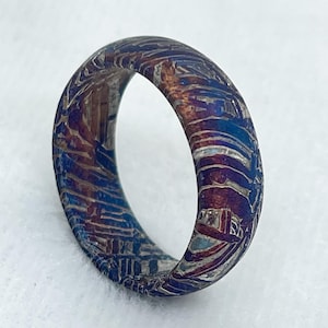 Könnte beinhalten: Ein Ring mit einem komplexen, mehrfarbigen Metallmuster mit tiefblauen, kupferfarbenen und silbernen Streifen. Die Oberfläche weist eine ausgeprägte, geätzte kristalline Textur auf, die sich um den gesamten Umfang des Rings erstreckt.