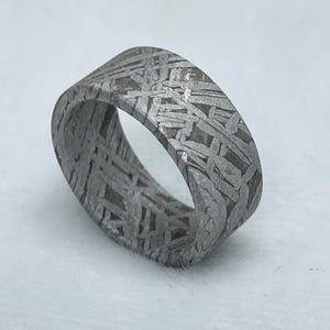 May include: A wide, patterned ring with a unique, textured design. The ring is made of a gray metal with a cross-hatched pattern across the surface. The ring's design is consistent throughout the band.