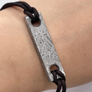 May include: A rectangular, silver-colored bracelet charm with a unique patterned surface is strung on a dark brown cord. The charm has two circular holes for the cord to pass through. The bracelet is worn on a wrist.