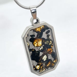 May include: A rectangular pendant necklace featuring a silver-toned metal frame containing embedded dark and golden-brown mineral fragments. The pendant hangs from a matching silver-toned snake chain, showcasing a clear, polished finish over the stones.