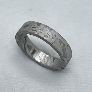 Puede incluir: Un anillo plateado con un patrón grabado detallado. El anillo tiene una banda plana y una superficie texturizada. El intrincado diseño cubre toda la banda, creando un aspecto único y artístico. El anillo se muestra sobre una superficie blanca.