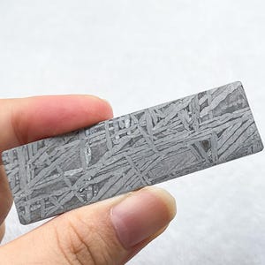 May include: A rectangular piece of iron meteorite featuring a distinct Widmanstätten pattern of intersecting metallic bands in shades of gray and silver, displaying a natural crystalline structure across its flat, polished surface.