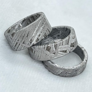 Puede incluir: Tres anillos fabricados en material metálico gris con un patrón de Widmanstätten distintivo de líneas entrecruzadas y formas geométricas en toda la superficie. Las bandas tienen un perfil plano y un acabado mate texturizado en todo el conjunto.