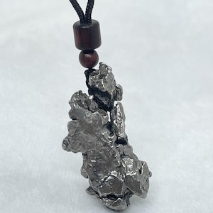 May include: A metallic, silver-toned pendant with a jagged, irregular, and textured surface. The pendant is suspended from a black cord featuring two dark brown, cylindrical and spherical wooden beads positioned above the metal piece.