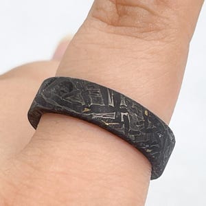 Peut inclure: Une bague gris foncé avec une surface mate et texturée présentant un motif géométrique complexe de lignes entrecroisées et de petites taches métalliques. L'anneau a un profil plat et une largeur constante, offrant un aspect complexe et patiné.