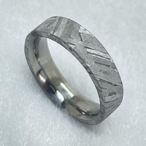 Puede incluir: Un anillo de metal en tono plateado que presenta un patrón de Widmanstätten distintivo en su superficie exterior. La banda tiene un perfil plano con un interior pulido y un acabado exterior texturizado que muestra líneas geométricas.