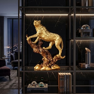 May include: A gold leopard sculpture on a textured, brown tree branch. The decorative figurine is displayed on a black shelf with other decor items. The leopard is in a climbing pose, with its front paws on the branch.