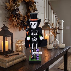 May include: A skeleton nutcracker wearing a black top hat with an orange band and a black cape. The figure holds a staff topped with a bat and features a sign at its base that reads Ghost Town. It has white bone details on a black body.