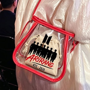 May include: A clear, red-trimmed crossbody bag with a red strap. Inside, a small beige drawstring bag features a black silhouette of people and the word "ARIRANG" with "BTS - 2026" below. The bag is worn over a white blouse.