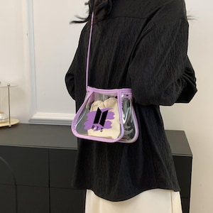 May include: A clear, small crossbody bag with a lavender trim and strap. The bag features a white interior pouch and a black and purple logo that reads "BTS". The bag is worn over a black top.
