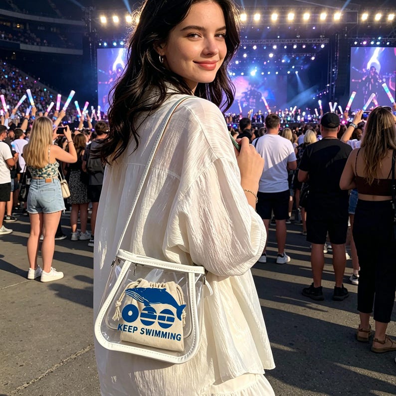 May include: A clear crossbody bag with a white trim and a drawstring pouch inside. The pouch features a blue whale graphic and the text "KEEP SWIMMING". The bag is worn over a white outfit.