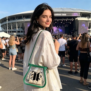 May include: A clear crossbody bag with green trim and a beige pouch featuring a black and white BTS logo and the word "BTS". The bag is worn over a white shirt. A crowd of people is in the background.