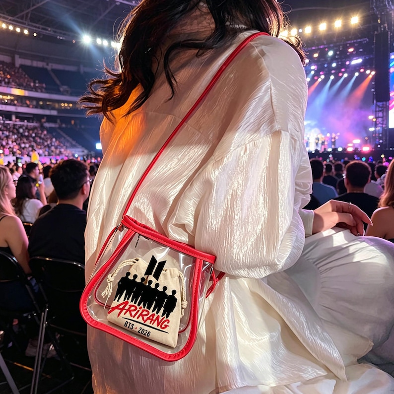 May include: A clear, red-trimmed crossbody bag with a red strap. Inside, a small beige drawstring bag features a black silhouette of people and the word "ARIRANG" with "BTS - 2026" below. The bag is worn over a white blouse.