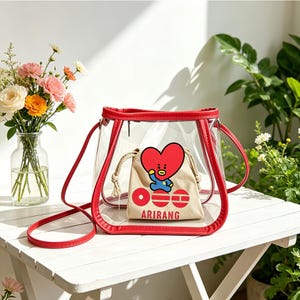 May include: A clear, red-trimmed handbag with a matching shoulder strap. Inside, a small beige drawstring bag features a heart-shaped character and the word "ARIRANG". The bag sits on a white table with a vase of flowers.