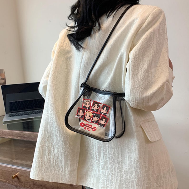 May include: Clear crossbody bag with black trim and a black strap. The bag features a graphic of seven cartoon figures with the text "BTS" in red and white. The bag is worn over a white blazer.