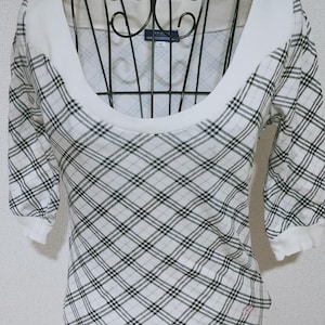 May include: A white and black checked top with a scoop neckline and short sleeves. The top is displayed on a black metal mannequin. The fabric has a diamond pattern. The top has a white collar and white trim on the sleeves and bottom.