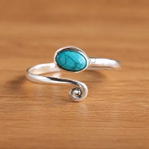 May include: A silver-toned ring with a turquoise oval stone. The ring has a swirling design on one end and a smooth band that wraps around the finger. The turquoise stone is a bright, vibrant blue-green color.