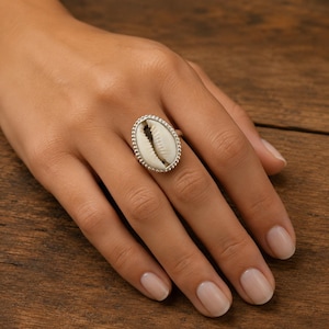 Può includere: Anello in argento di forma ovale con una conchiglia bianca naturale. L'anello ha un bordo con perline. L'anello è mostrato su una mano con smalto neutro, su uno sfondo di legno. Design semplice ed elegante.