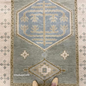 May include: A rectangular area rug with a central geometric design in shades of blue, beige, and green. The rug features a central diamond shape with floral motifs and a border with repeating cross patterns. The rug is on a light beige background.