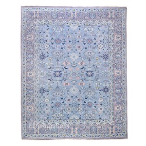 May include: A rectangular area rug featuring a light blue field with a repeating floral and vine pattern in shades of navy, soft pink, and cream. The rug has a wide, intricate border in a muted lavender-grey tone with matching floral motifs.