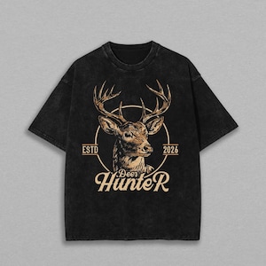 May include: A black short-sleeve t-shirt with a washed-out texture. The center features a tan graphic of a deer head inside a circle, with the text ESTD 2026 on the sides and the word Deer Hunter printed in a stylized font below the circle.